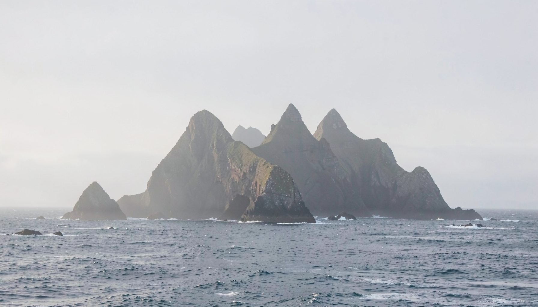 Misty view of Skellig Michael's jagged silhouette against a pale grey Atlantic horizon, with gentle waves lapping at its rocky base in soft morning light