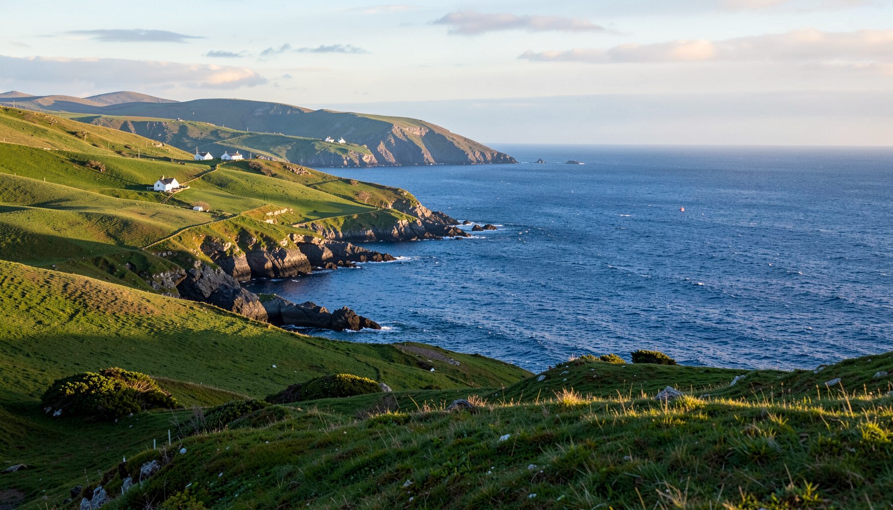 Rolling green hills meeting the deep blue Atlantic along the Skellig Coast, with soft golden light and scattered white cottages near the shoreline.