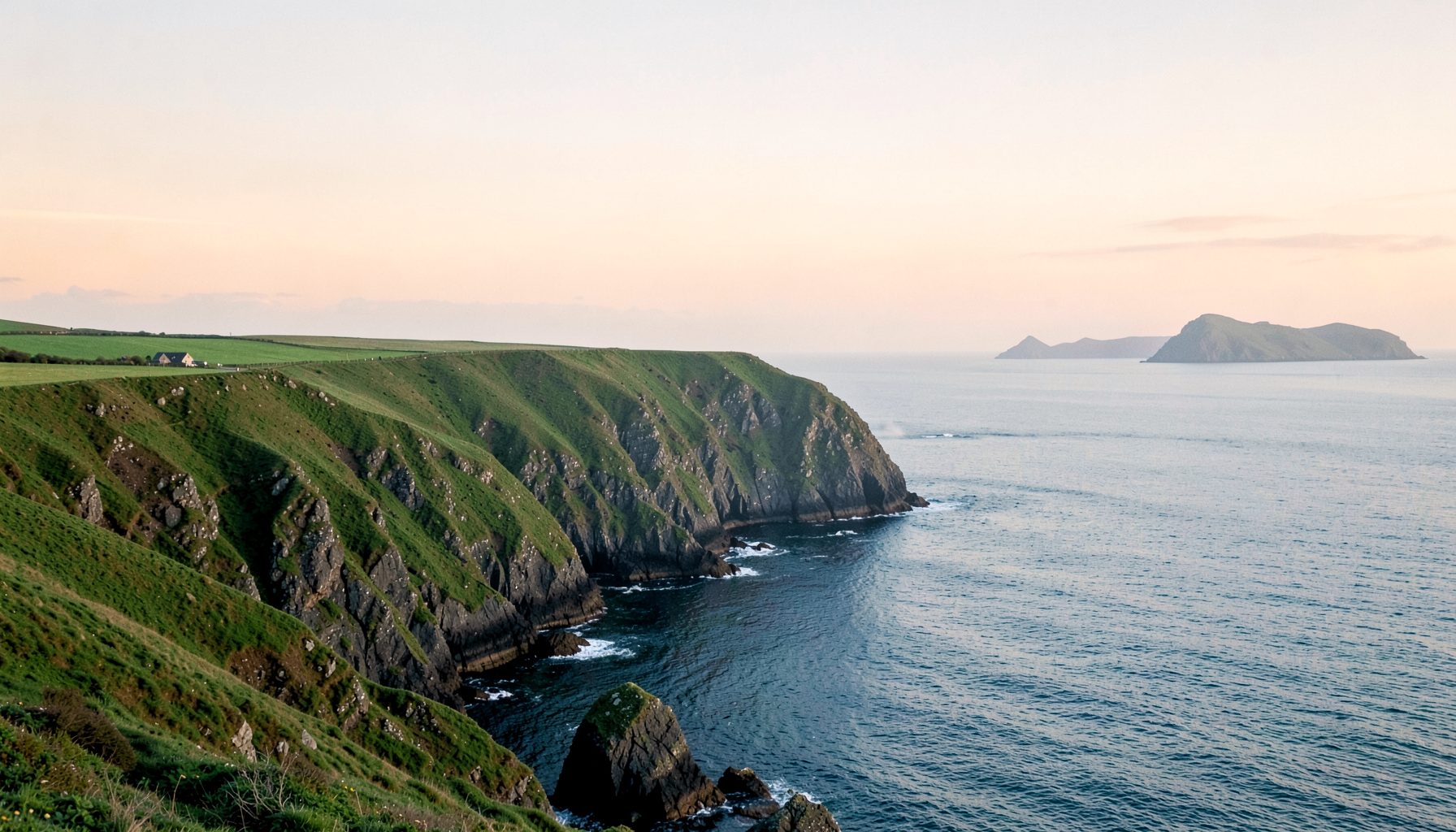 A wide sweep of rugged green coastal cliffs meeting a calm blue-grey sea under a soft peach-tinted sky, with distant rocky island peaks on the horizon, evoking a quiet windswept Irish coastline