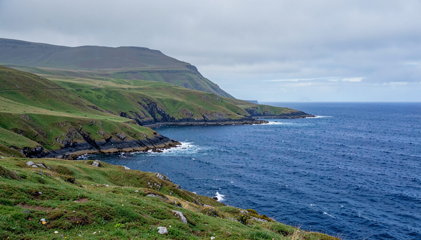 A sweeping view of the Skellig Coast on the Iveragh Peninsula, with green rolling hills meeting the deep blue Atlantic under a soft grey Irish sky.