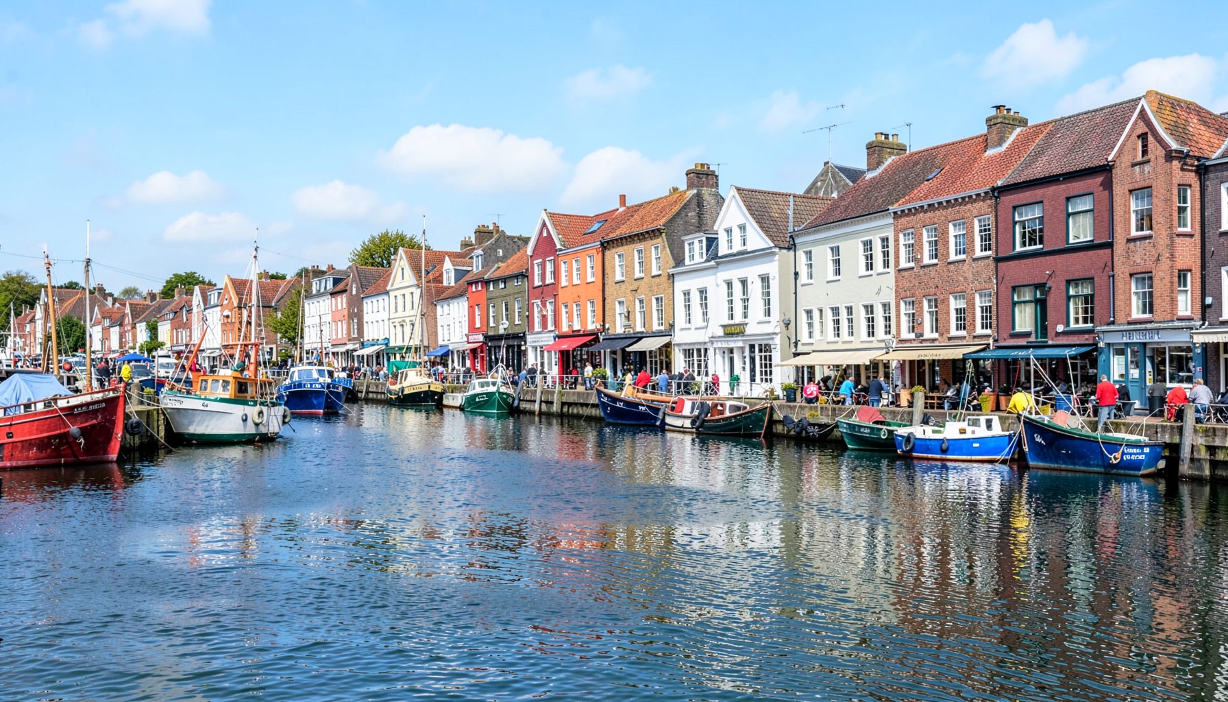 A colourful row of houses and pubs along a waterfront, with fishing boats moored in a sheltered harbour under a bright sky