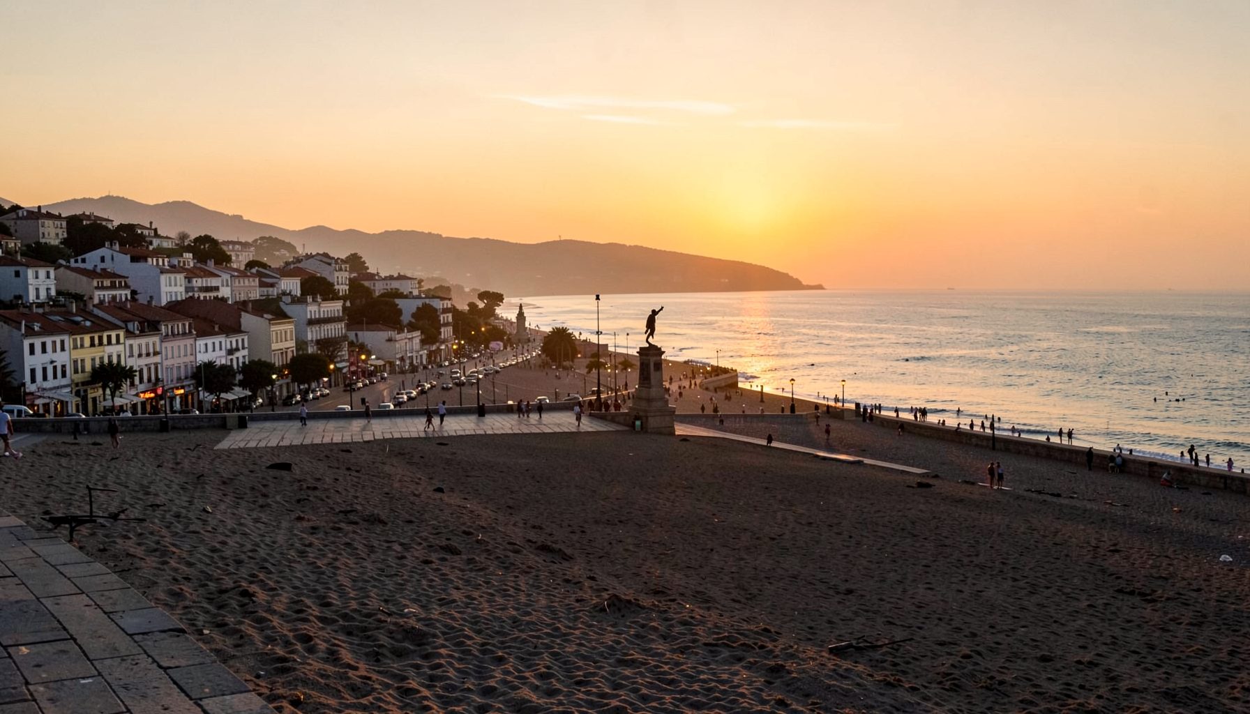 A seaside town with a long sandy beach, a promenade, and a statue silhouetted against a golden sunset over the water